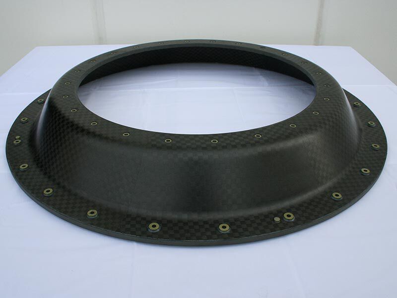 Aspect angle looking at the smallsat launch adapter structural ring. There are machined  holes with metallic inserts along the bottom and top flat edges of the ring., The surface is a carbon fiber weave with a matte finish. The ring sits upon a white table with a neutral background.